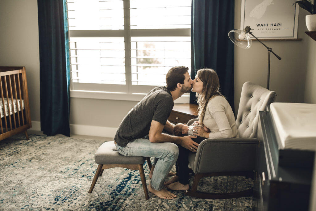 Mom and dad kissing with newborn in their arms sitting in the nursery