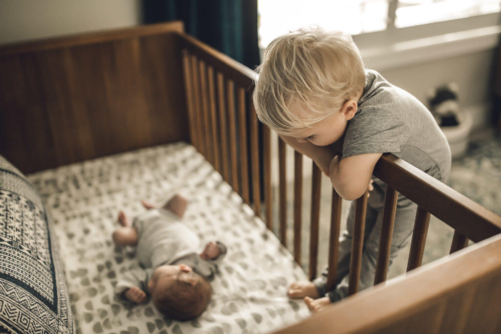 Blonde-haired toddler looking over into sleeping baby's crib