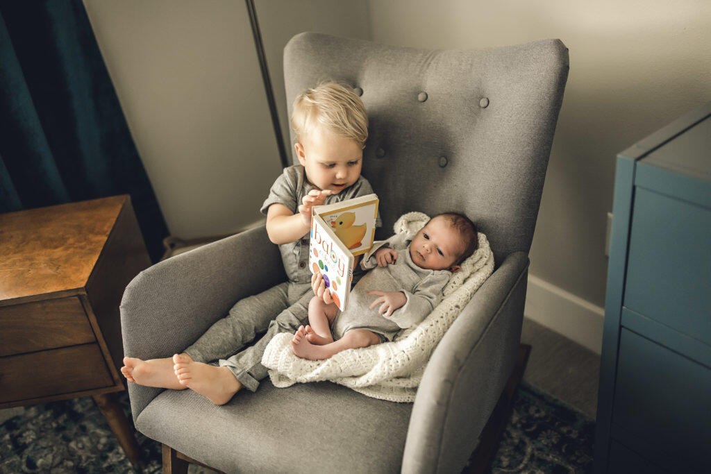 Blonde-haired toddler reading to newborn brother in nursery rocker