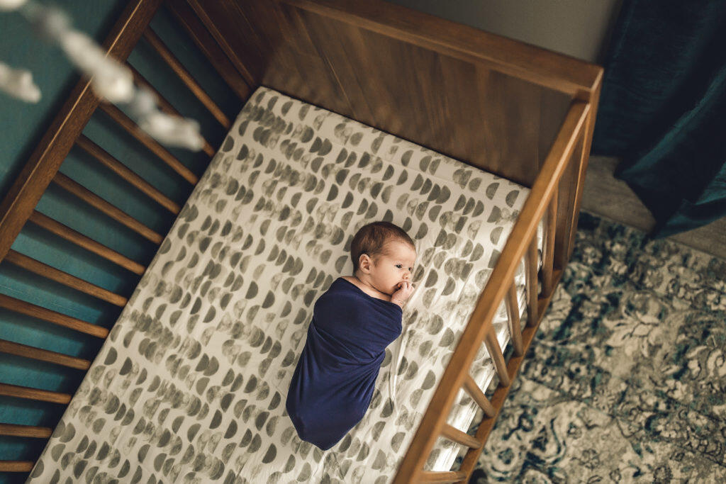 Newborn swaddled in navy swaddle laying in warm oak crib with half moon sheets